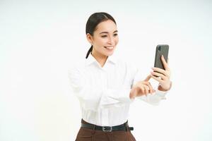 hermosa sonriente mujer de negocios toma selfie en móvil teléfono, hace un foto en teléfono inteligente, registros video, soportes terminado blanco antecedentes