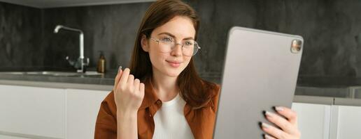 Portrait of beautiful woman in glasses, looking at digital tablet, reading on gadget, taking selfie on device photo