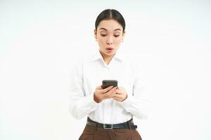 Portrait of asian businesswoman looks at smartphone with surprised face, amazing news on mobile phone message, standing over white background photo