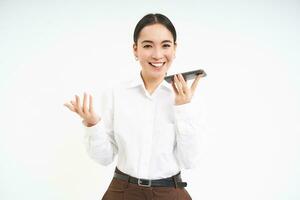 sonriente hembra vendedora, asiático mujer gerente negociaciones en altavoz teléfono, sostiene Teléfono móvil cerca boca y traduce su discurso, blanco antecedentes foto