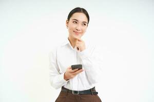 Asian business woman, young manager holds smartphone and thinks, looks thoughtful aside, makes decision, stands over white background photo