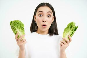 Portrait of woman with surprised face, holding cabbage, lettuce leaves and looks amazed, growing vegetables in own garden, white background photo