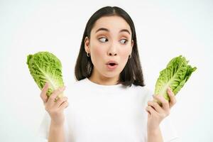 Portrait of woman with surprised face, holding cabbage, lettuce leaves and looks amazed, growing vegetables in own garden, white background photo
