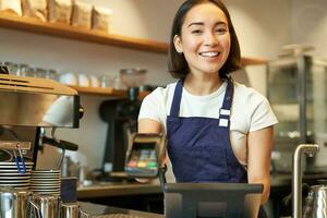 pequeño negocio y gente. sonriente asiático mujer barista, trabajando en cafetería, dando pos Terminal, crédito tarjeta máquina, Procesando orden foto