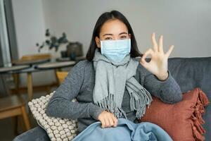 Covid-19 and health concept. Young asian woman in medical face mask, feels sick and unwell, catching flu, protecting others from influenza, sitting in living room covered with blanket photo