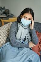 Covid-19 and health concept. Young asian woman in medical face mask, feels sick and unwell, catching flu, protecting others from influenza, sitting in living room covered with blanket photo