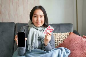 Smiling korean girl shows smartphone screen, medication in hands, feeling sick and staying at home, using online GP doctor app while on sick leave photo