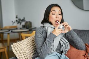 Ill woman using spray from sore throat, treating her cold or flu, staying at home on sick leave, sitting on sofa in living room photo