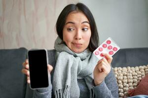 Happy asian woman catching cold, showing smartphone screen and pills, recommending online doctor app, GP or medical clinic on phone, holding drugs, staying at home with flu photo