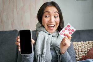 Happy asian woman catching cold, showing smartphone screen and pills, recommending online doctor app, GP or medical clinic on phone, holding drugs, staying at home with flu photo