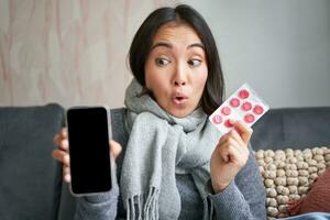 Happy asian woman catching cold, showing smartphone screen and pills, recommending online doctor app, GP or medical clinic on phone, holding drugs, staying at home with flu photo