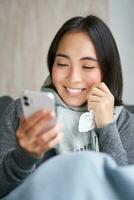 Portrait of korean woman feels sick, holding smartphone, calling doctor gp to get presctiption, caught cold, staying at home, using mobile phone photo