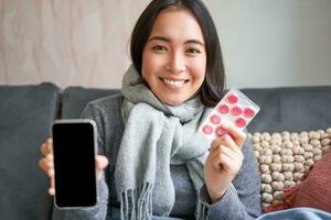 Smiling korean girl shows smartphone screen, medication in hands, feeling sick and staying at home, using online GP doctor app while on sick leave photo