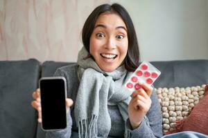 Smiling korean girl shows smartphone screen, medication in hands, feeling sick and staying at home, using online GP doctor app while on sick leave photo