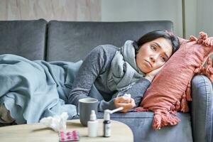 Sick sad korean woman lying on sofa, feeling unwell, catching cold, flu and temperature, looking upset, taking medication photo