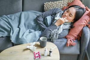 Sick sad korean woman lying on sofa, feeling unwell, catching cold, flu and temperature, looking upset, taking medication photo
