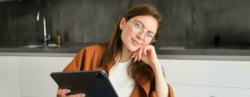 Portrait of busy young woman, self-employed entrepreneur sitting at home with digital tablet, reading documents. Student working on project remotely, holding gadget, studying photo