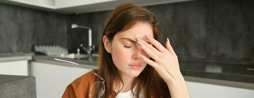 Close up portrait of brunette woman touching head, feeling unwell, has headache or migraine, sitting in kitchen with dizziness. photo