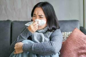 Close up of korean girl sneezing in napking, sitting at home in scarf, feeling unwell, concept of winter, heating cost and people photo