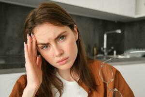 Close up portrait of tired young woman, wife sitting in kitchen and touching head, has headache, massaging temple and frowning from painful migraine photo