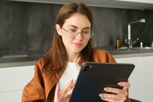 Portrait of busy young woman, self-employed entrepreneur sitting at home with digital tablet, reading documents. Student working on project remotely, holding gadget, studying photo