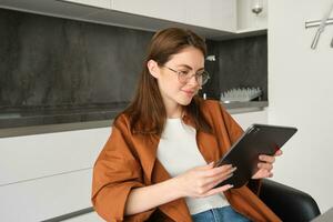 Portrait of young brunette woman in glasses, sitting in her kitchen with digital tablet, reading on gadget, watching videos online, working on remote from her home photo