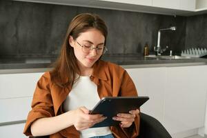 Portrait of brunette woman in glasses, sitting in kitchen at home, holding digital tablet, reading on gadget photo