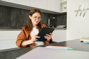 Portrait of young brunette woman in glasses, sitting in her kitchen with digital tablet, reading on gadget, watching videos online, working on remote from her home photo