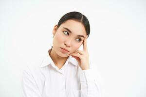 Portrait of annoyed, bored korean woman roll eyes, looks aside with bothered, tired face expression, stands over white background photo