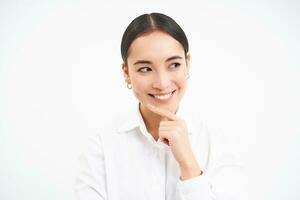 Successful asian woman, business professional, smiling with intrigued face, thinking, looking thoughtful aside, standing over white background photo