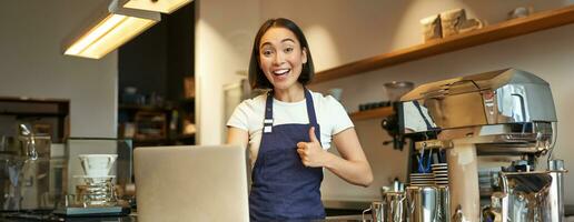 Portrait of asian girl in apron, barista in coffee shop, shows thumbs up, approve or recommend smth, working in cafe, smiling happy photo