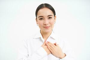 Portrait of asian businesswoman, looks with care, holds hands on heart, stands over white background photo