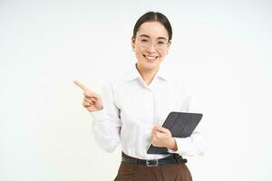 Young professional, businesswoman in glasses holding digital tablet, pointing finger left, showing business advertisement, standing over white background photo