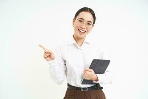 Young professional, businesswoman in glasses holding digital tablet, pointing finger left, showing business advertisement, standing over white background photo