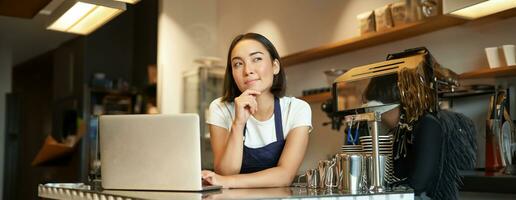 Portrait of girl barista with laptop, standing at counter in coffee shop and thinking, looking curious and thinking photo