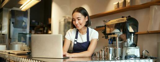 Portrait of smiling asian barista, cafe owner entrepreneur, working on laptop, processing orders on computer, standing behind counter photo