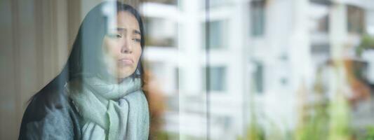 Portrait of sad ill korean woman looking out of the window, staying at home due to coronavirus, being sick and cant leave house photo