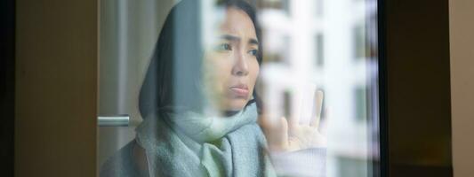 Portrait of sad ill korean woman looking out of the window, staying at home due to coronavirus, being sick and cant leave house photo