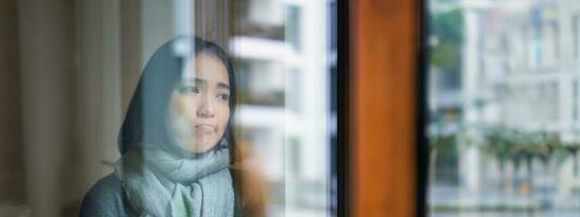 Portrait of sad ill korean woman looking out of the window, staying at home due to coronavirus, being sick and cant leave house photo