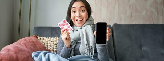 en línea doctores y cuidado de la salud. sonriente coreano mujer muestra móvil teléfono pantalla y medicamento, atrapando frío, quedarse a hogar y recomendando teléfono inteligente gp foto