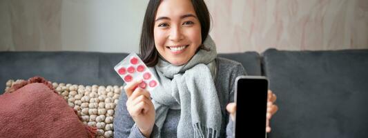 Smiling korean girl shows smartphone screen, medication in hands, feeling sick and staying at home, using online GP doctor app while on sick leave photo