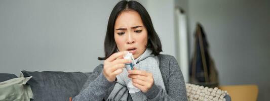 Ill korean woman looking at thermometer with concerned face, catching cold, having fever, having covid, staying at home on sick leave photo