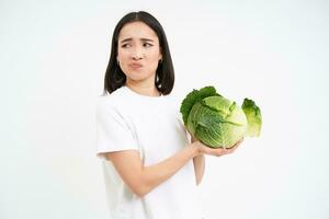 Asian woman looks with disgust and dislike at cabbage, hates vegetables, isolated on white background photo