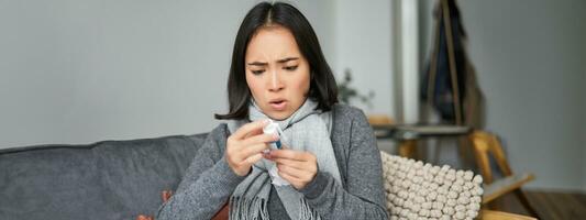 Ill korean woman looking at thermometer with concerned face, catching cold, having fever, having covid, staying at home on sick leave photo