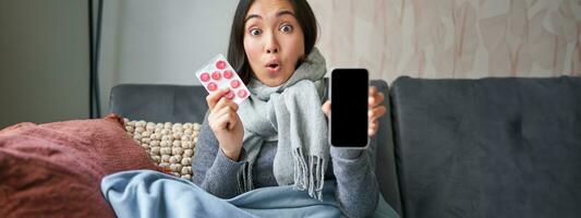 Online doctors and healthcare. Smiling korean woman shows mobile phone screen and medication, catching cold, staying at home and recommending smartphone GP photo