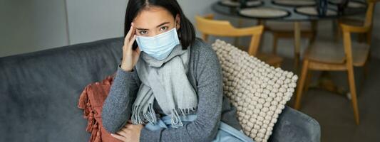 Woman in medical face mask has covid-19, stays at home in personal protective equipment, catching flu, sitting in living room photo