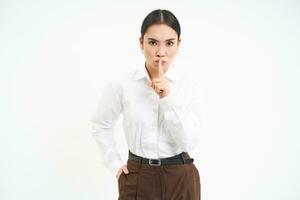 Strict and serious businesswoman shushing, puts finger on lips, tells to be quiet, hush gesture, stands over white background photo