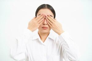 Asian woman entrepreneur, businesswoman shuts her eyes with hands, stands blindfolded against white studio background photo