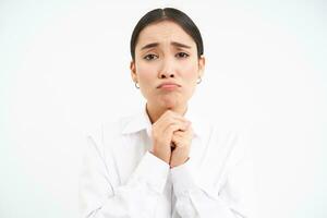 Upset young saleswoman, asian woman holds hands in pray, begs you, asks for favour with sad face, pleading, standing over white background photo