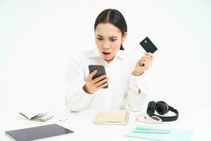 Young woman student, sits with smartphone and credit card, looks shocked at her bank account on mobile phone app, sitting frustrated over white background photo